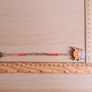 Pu&ograve; includere: Un ciondolo di perline con un personaggio di mucca Highland arancione chiaro attaccato a una chiusura a moschettone color argento. Il ciondolo misura circa 19 cm di lunghezza, come indicato dai righelli in legno visibili sul lato e in basso.