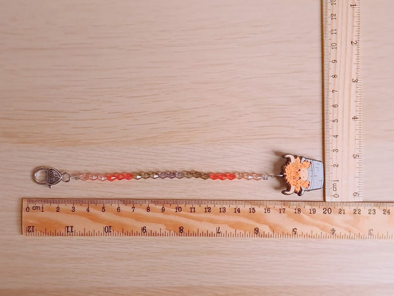 Pu&ograve; includere: Un ciondolo di perline con un personaggio di mucca Highland arancione chiaro attaccato a una chiusura a moschettone color argento. Il ciondolo misura circa 19 cm di lunghezza, come indicato dai righelli in legno visibili sul lato e in basso.