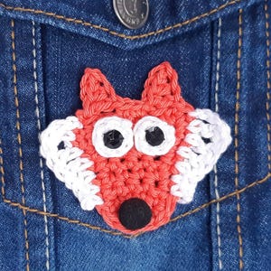Puede incluir: Primer plano de un bolsillo de chaqueta vaquera con un aplique de zorro de ganchillo. El zorro es rojo con detalles blancos, ojos negros y nariz negra. La chaqueta tiene un botón y costuras visibles.