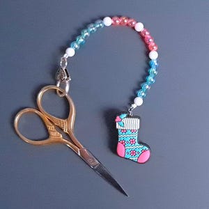 May include: Gold-colored embroidery scissors with a decorative handle, paired with a beaded charm featuring a Christmas stocking design in pink, blue, and white. The charm is attached to the scissors with a silver-colored clasp.