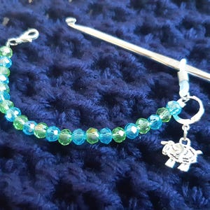 May include: A blue and green crystal bead bracelet with a silver charm of a sheep attached to a silver crochet hook.