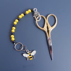 May include: A pair of small gold scissors with a black and yellow beaded chain attached to a bee charm.