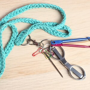 Pode incluir: Um cord&atilde;o de croch&ecirc; turquesa com um chaveiro prateado e um conjunto de agulhas de croch&ecirc; coloridas e um pequeno par de tesouras.