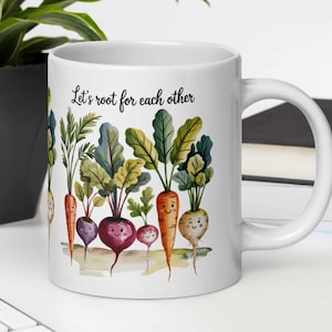 Peut inclure: Une tasse en céramique blanche avec une illustration colorée de carottes et de navets avec des visages souriants. La tasse porte l'inscription "Let's root for each other".