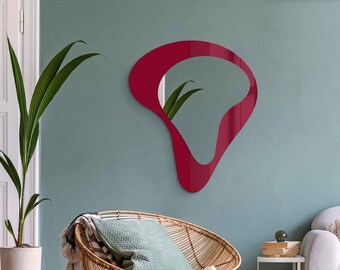 May include: A red abstract shaped wall mirror with a wavy design hangs on a light blue wall above a wicker chair with a green cushion and a striped throw blanket.