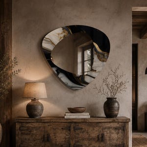 May include: An abstract-shaped wall mirror with a black, white, and gold marbled design hangs above a wooden cabinet. A lamp with a beige shade sits on the cabinet, and a large vase with a plant is on the left. The room has a rustic, natural aesthetic.