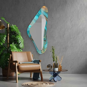 May include: A teal and gold abstract patterned mirror with a unique, asymmetrical shape hangs on a gray wall. A brown leather armchair with a blue throw sits in front of the mirror. A small glass table with a blue base sits in front of the chair.