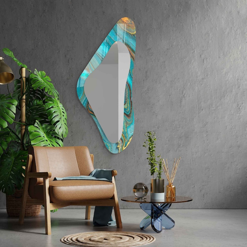 May include: A teal and gold abstract patterned mirror with a unique, asymmetrical shape hangs on a gray wall. A brown leather armchair with a blue throw sits in front of the mirror. A small glass table with a blue base sits in front of the chair.