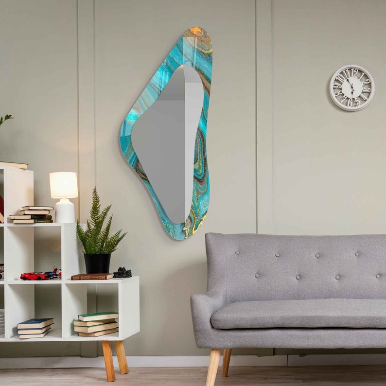 May include: A teal and gold abstract patterned wall mirror with an irregular, organic shape. The mirror is hanging on a white wall in a living room setting.