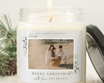 May include: A lit, clear glass jar candle with a black lid, featuring a family photo and the text "MERRY CHRISTMAS with love, THE BROWNS." The candle is surrounded by a pine branch and a pine cone, suggesting a holiday theme.