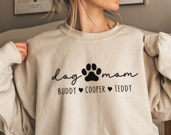 Könnte beinhalten: Ein beiger Pullover mit dem Text "dog mom" und einer Pfotenabdruckgrafik. Der Text "Buddy Cooper Teddy" ist unter dem Pfotenabdruck gedruckt.