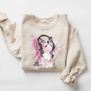 May include: A beige sweatshirt with a graphic print of a cat wearing headphones. The cat is looking up and has pink and blue watercolor accents around it.