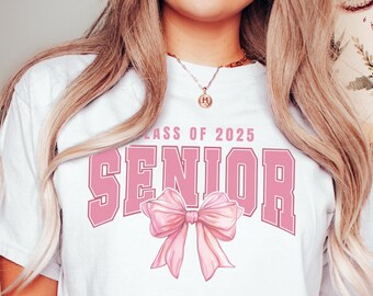 Puede incluir: Camiseta blanca con el texto "Class of 2025 Senior" en letras rosas y un lazo rosa.