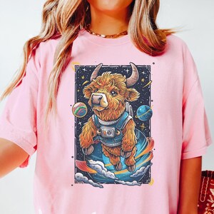 May include: A pink t-shirt with a white graphic print of a brown highland cow wearing a space suit and floating in space. The cow is surrounded by stars and planets. The text "Blossom Shirt" is printed below the image.