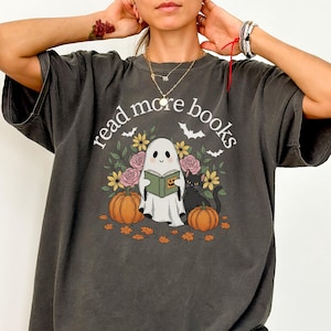 May include: Dark grey t-shirt featuring the text "read more books" above a graphic of a ghost reading a book, pumpkins, flowers, bats, and a black cat. The shirt has a relaxed fit.