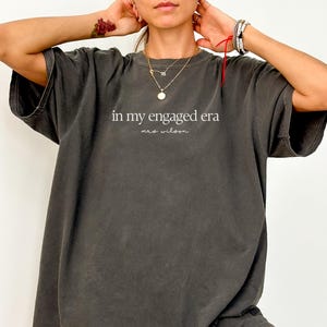 May include: An oversized t-shirt in a dark grey colour with the text "in my engaged era" and "mrs. wilson" in white lettering.