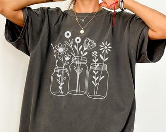 May include: Dark grey t-shirt featuring a white illustration of flowers in mason jars. The shirt has a loose fit and short sleeves. The text "COLOR: PEPPER" is visible at the bottom of the image.