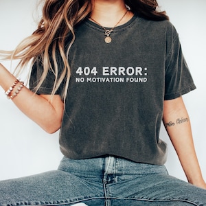 May include: A dark gray t-shirt with the text "404 ERROR: NO MOTIVATION FOUND" printed in white. The shirt is worn by a person sitting down, wearing blue jeans and white sneakers.
