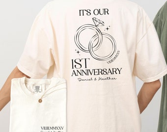 May include: Ivory t-shirt with a graphic of two intertwined rings with a diamond, the text "It's Our 1st Anniversary" and the names "Daniel & Heather" in black.
