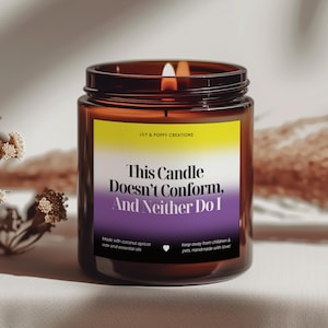 Puede incluir: Una vela de vidrio marrón con una etiqueta amarilla, morada y blanca que dice "This Candle Doesn't Conform, And Neither Do I". La vela está encendida y tiene un símbolo de corazón en la etiqueta.