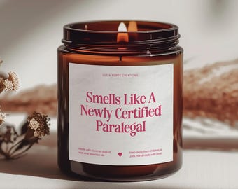 May include: A brown glass candle with a white label that reads "Smells Like A Newly Certified Paralegal". The candle is lit and has a pink heart icon on the label.