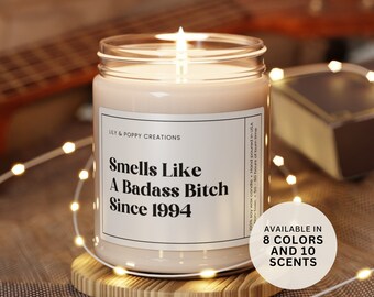 May include: A glass candle jar with a white label that reads "Smells Like A Badass Bitch Since 1994". The candle is lit and the jar is sitting on a wooden coaster. The text "AVAILABLE IN 8 COLORS AND 10 SCENTS" is on the label.