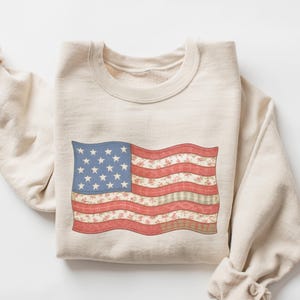 May include: Sand-colored sweatshirt featuring a distressed American flag design. The flag has a blue field with white stars and red and floral-patterned stripes. The text "COLOR: SAND" is at the bottom of the image.