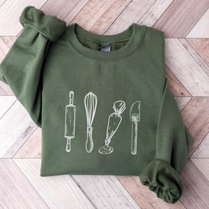 Könnte beinhalten: Ein grünes Sweatshirt mit einer weißen Grafik von Backutensilien, darunter ein Nudelholz, ein Schneebesen, ein Spritzbeutel und ein Spatel.