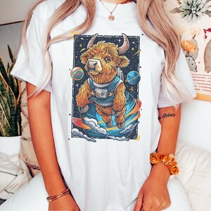 May include: White t-shirt with a graphic of a brown and white furry cow wearing a space suit and floating in space. The cow is surrounded by planets and stars.