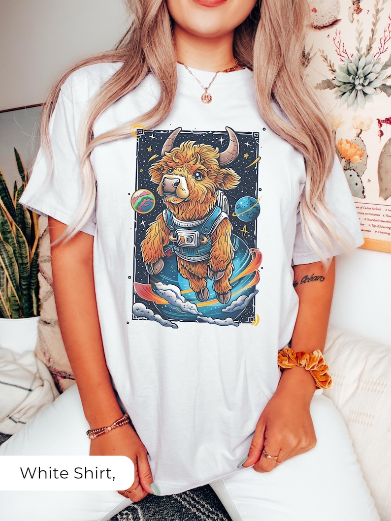 May include: White t-shirt with a graphic of a brown and white furry cow wearing a space suit and floating in space. The cow is surrounded by planets and stars.