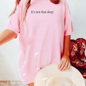 May include: An oversized pink t-shirt with the text "it's not that deep" printed on the front. The shirt is being worn by a person holding a straw hat. The colour of the shirt is called "blossom".