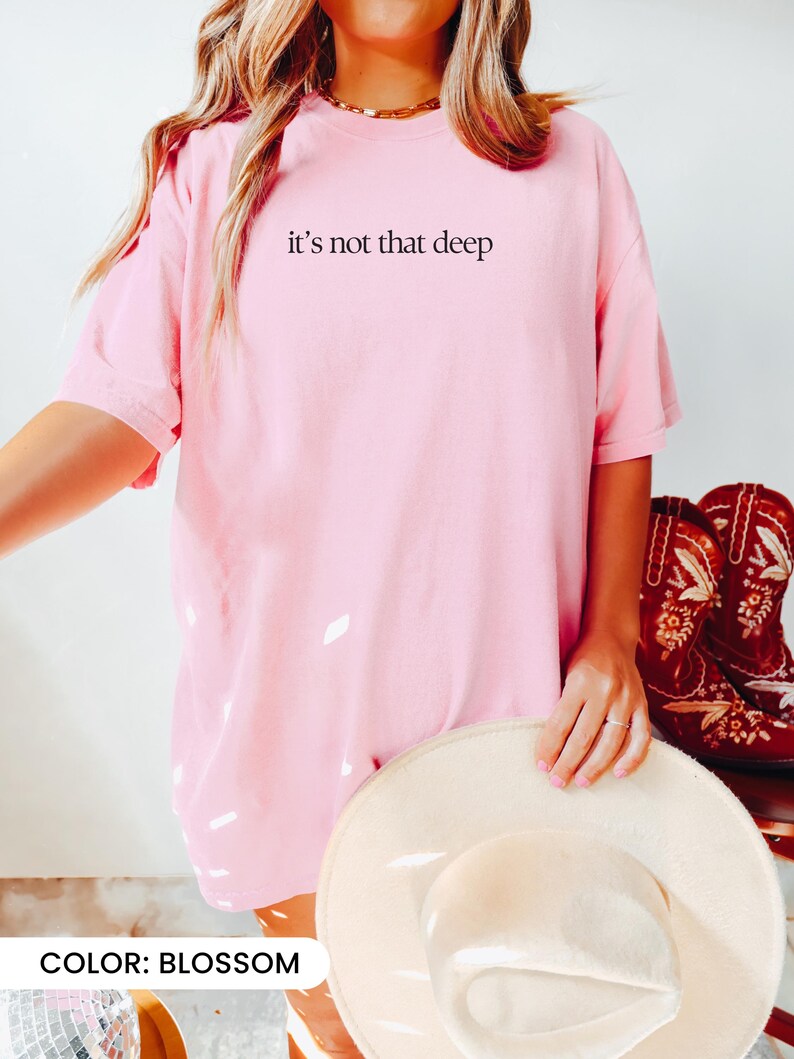 May include: An oversized pink t-shirt with the text "it's not that deep" printed on the front. The shirt is being worn by a person holding a straw hat. The colour of the shirt is called "blossom".