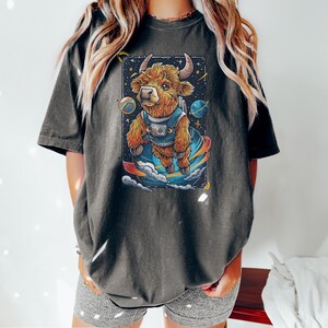 May include: A dark gray t-shirt with a cartoon illustration of a brown highland cow wearing a space suit and floating in space. The cow is surrounded by stars and planets.