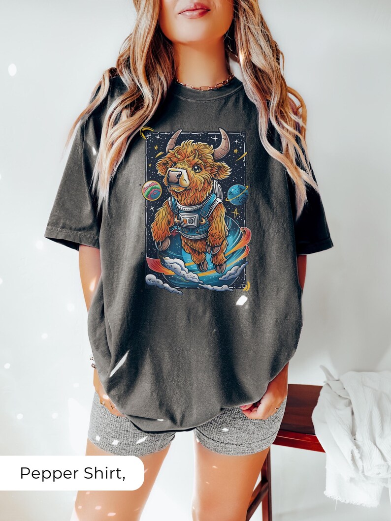 May include: A dark gray t-shirt with a cartoon illustration of a brown highland cow wearing a space suit and floating in space. The cow is surrounded by stars and planets.