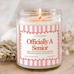 May include: A clear glass jar candle with a lit wick, featuring a label with pink vertical stripes and a scalloped border. The text reads: Lily & Poppy Creations, Officially A Senior, Smell that? That's twelve years of homework and one last victory lap. Net weight 9oz.