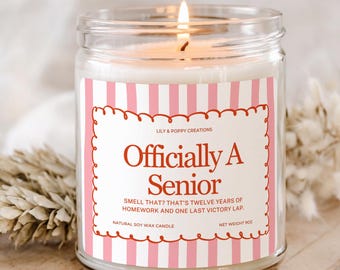 Puede incluir: Una vela en frasco de vidrio transparente con una mecha encendida, con una etiqueta de rayas verticales rosas y borde festoneado. El texto dice: Lily & Poppy Creations, Officially A Senior, Smell that? That's twelve years of homework and one last victory lap. Peso neto 9oz.
