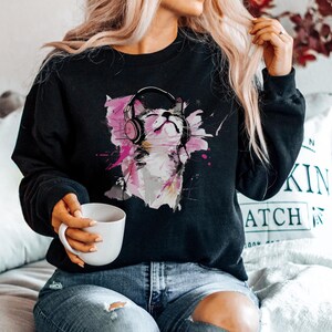 May include: Black sweatshirt with a colorful graphic of a cat wearing headphones. The cat is surrounded by pink and purple paint splatters.