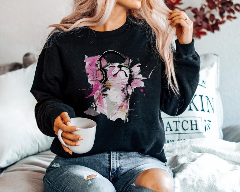 May include: Black sweatshirt with a colorful graphic of a cat wearing headphones. The cat is surrounded by pink and purple paint splatters.