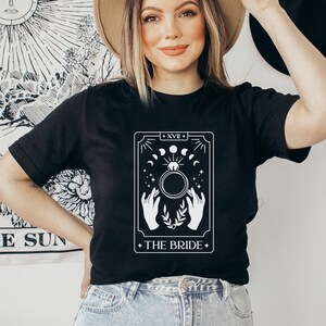 Pode incluir: Camiseta preta com um design de carta de tar&ocirc; branco. A carta &eacute; rotulada como "A Noiva" e apresenta um anel com uma lua e estrelas ao redor.