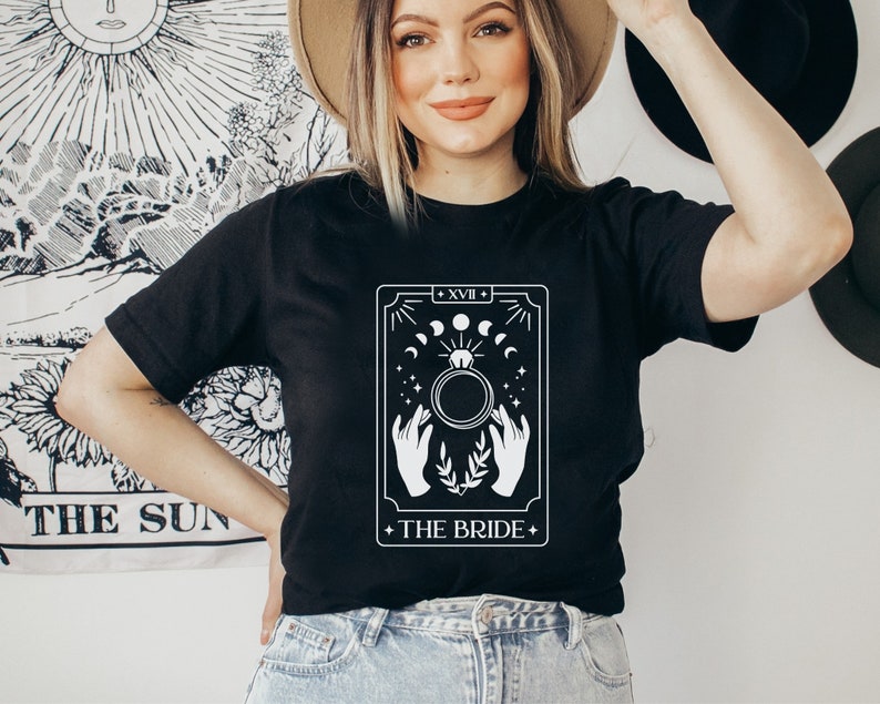 Pode incluir: Camiseta preta com um design de carta de tar&ocirc; branco. A carta &eacute; rotulada como "A Noiva" e apresenta um anel com uma lua e estrelas ao redor.