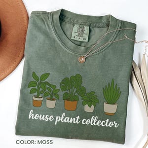 Op de afbeelding: Mosgroen t-shirt met een afbeelding van potplanten en de tekst "house plant collector". Het shirt heeft een ronde hals en korte mouwen. Een gouden ketting met een kleine hanger ligt op het shirt.