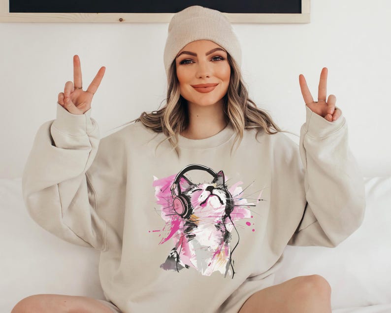 May include: A beige sweatshirt with a graphic print of a cat wearing headphones. The cat is drawn in a watercolor style with pink, gray, and black accents.