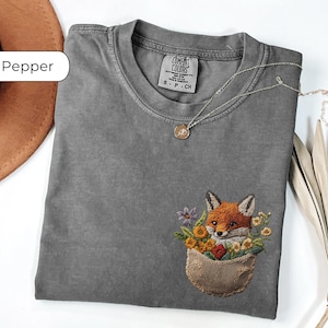 May include: A grey t-shirt with an embroidered fox in a pocket. The fox is surrounded by flowers and the text "Comfort Colors" is on the tag.