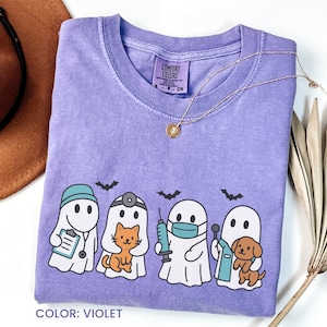 Puede incluir: Camiseta violeta Comfort Colors con un gráfico de cuatro personajes fantasma vestidos como profesionales médicos, cada uno con una mascota. El diseño incluye murciélagos y el texto "COLOR: VIOLET". Un collar dorado está drapeado sobre la camisa.