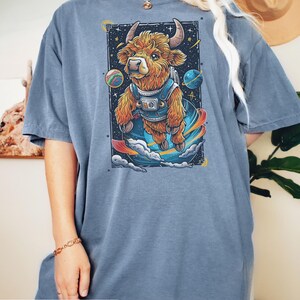 May include: A blue jean oversized t-shirt with a graphic of a brown and white highland cow wearing a space suit and floating in space. The cow is surrounded by planets and stars.