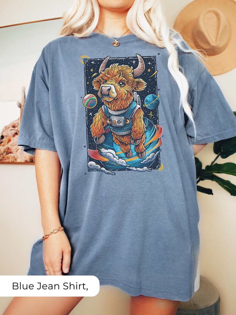 May include: A blue jean oversized t-shirt with a graphic of a brown and white highland cow wearing a space suit and floating in space. The cow is surrounded by planets and stars.