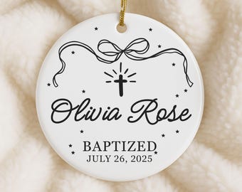 Może przedstawiać: Biała ceramiczna ozdoba z złotym sznurkiem do zawieszenia. Zawiera imię "Olivia Rose", słowo "BAPTIZED" i datę "26 lipca 2025" w czarnym skrypcie. Krzyż i gwiazdy zdobią projekt.