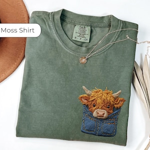 May include: A moss green t-shirt featuring an embroidered Highland cow design. The cow is brown and tan, set in a denim pocket. The shirt has a crew neck and short sleeves. A gold necklace is draped across the shirt.