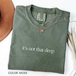 May include: A moss green t-shirt with the text "it's not that deep" in white. The shirt is made of a soft, comfortable material and has a relaxed fit.