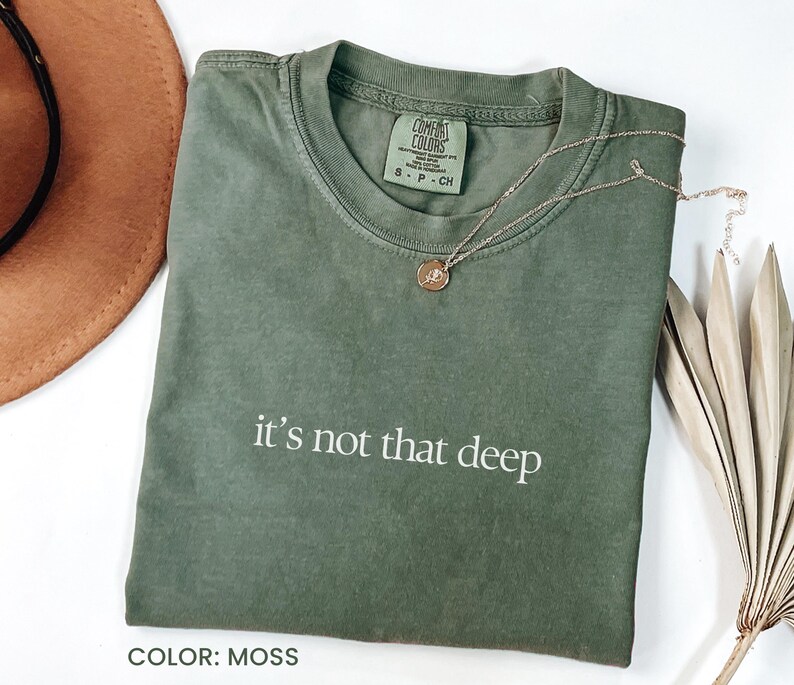 May include: A moss green t-shirt with the text "it's not that deep" in white. The shirt is made of a soft, comfortable material and has a relaxed fit.